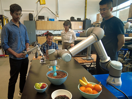 Students standing around a robotic arm that is assessing several bowls of fruit.