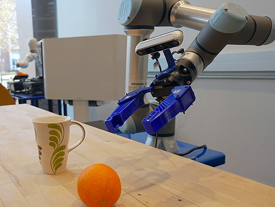 A robot arm reaching for a coffee cup and orange on the table in front of it.