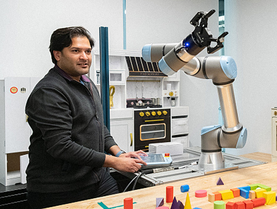 Abhinav Shrivastava standing at a table, on which a robot arm is mounted. There are assorted blocks and object on the tabletop for the arm to pick up.