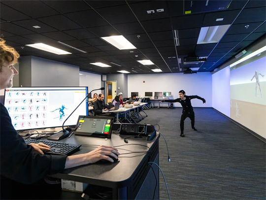 A classroom in which students are watching a student and professor working with a motion capture suit and software displaying various recorded poses.