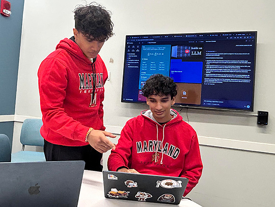 Two male students working at a laptop, with a large discplay screen behind them.