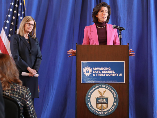 Two women in business attire on a stage, one is standing in front of the US flag, and the other is at a podium.