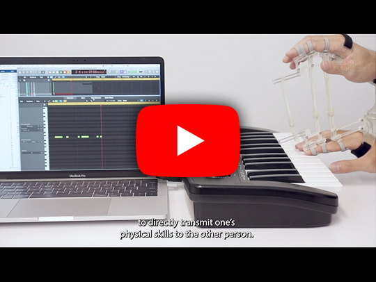 A still from a video with a red YouTube play button on it, showing hands wearing an exoskeleton, playing an electric piano keyboard. A monitor displays a recording of the hadn's actions. Click to play the video.