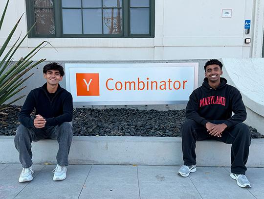 Two young men sitting outside in front of the Y Combinator sign