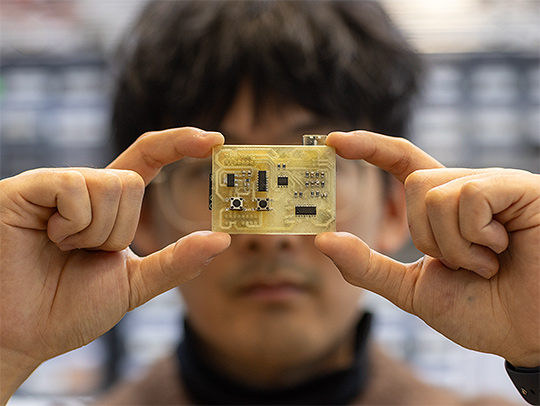 Closeup of a small circuit board held up in the hands of Zeyu Yan, who is seen out of focus behind it