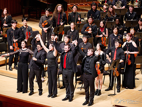 The Gamer Symphony Orchestra about to take a bow onstage after a concert. Everyone is dressed in black evening attire and most are holding instruments.
