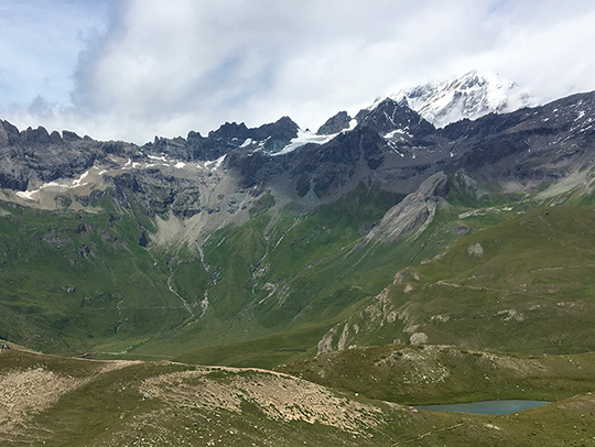 A landscape showing jagged peaks with green lower slopes, rolling hills with rivers running down them, a cloudy sky and a small lake.