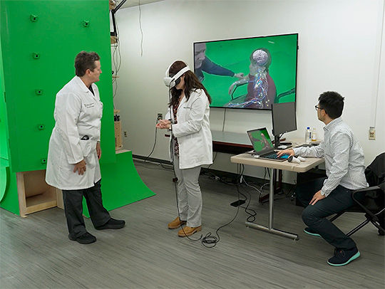 Three people in the holocamera suite: A female doctor in a lab coat, a female student in a lab coat wearing a VR headset, and a man sitting at a desk with a laptop. A large screen displays what the student is seeing via the headset. Three people in the holocamera suite: A female doctor in a lab coat, a female student in a lab coat wearing a VR headset, and a man sitting at a desk with a laptop. A large screen displays what the student is seeing via the headset.