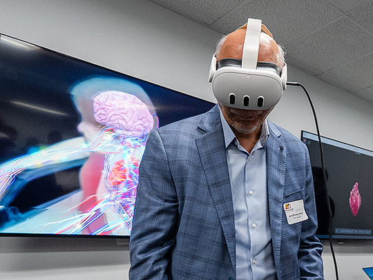 A middle-aged man wearing a VR headset. A large screen with a rendered environment is behind him.