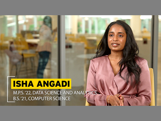 A video still of graduate student Isha Angadi. She is seated in the foreground. Her name and the program she is in is in the lower left. Behind her, a computer lab with someone in it can be seen in soft focus.