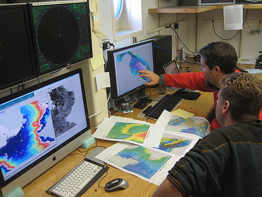 Co-lead scientists Jasper Konter and Matt Jackson at a desk covered in monitors and printed maps and, looking for a suitable seamount sampling site using data of collected by a research ship's sonar system. Credit: Val Finlayson.