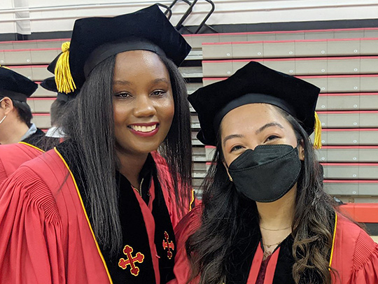 A Black Ph.D. graduate standing next to a fellow graduate. She is wearing Ph.D. regalia.