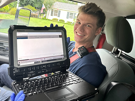 Kyle Zibell sitting in the front seat of a car. He is holding out a heavy-duty laptop displaying a software interface and wearing latex gloves.