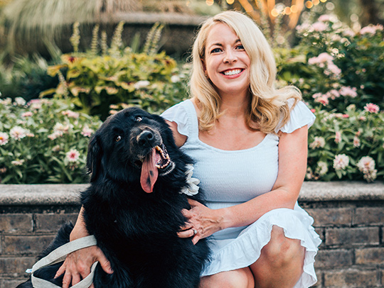 Lynsey White kneeling beside her large black dog. A fountain and flowers are in the background. Lynsey White kneeling beside her large black dog. A fountain and flowers are in the background.