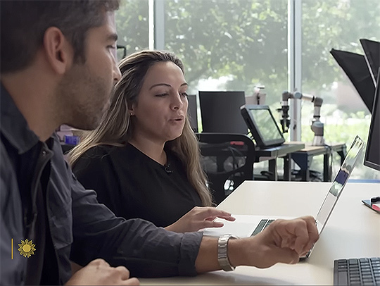 A video still of Marina Molina at a desk, pointing out something on her laptop screen to the person next to her. Credit: CBS.