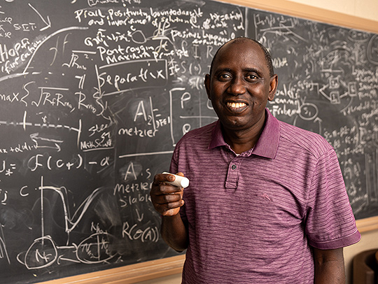 Abba Gumel with a large piece of chalk in his hand, standing in front of ablackboard filled with equations. Credit: Mark Sherwood.