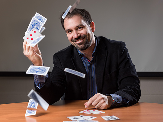 Matt Baker seated at a table with playing cards flying from his hand