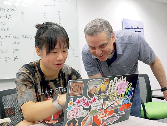 UMD mathematics Lecturer Todd Rowland leaning in to look a female high school student's laptop screen. Her laptop lid is covered with colorful stickers. Behind the pair is a glassboard full of equations. Credit: Mark Sherwood.