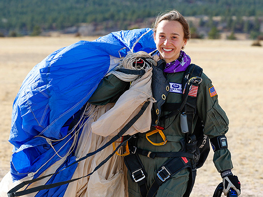 Maddie Fischer wearing a dark green US Air Force flight suit, walking with a gathered-up blue parachute slung over her shoulder.