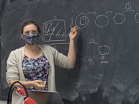 Marie Brodsky at a chalkboard with figures on it, teaching math.