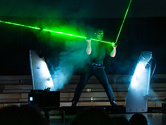 Magician Jason Latimer a darkened, foggy stage performing a trick with green lasters.