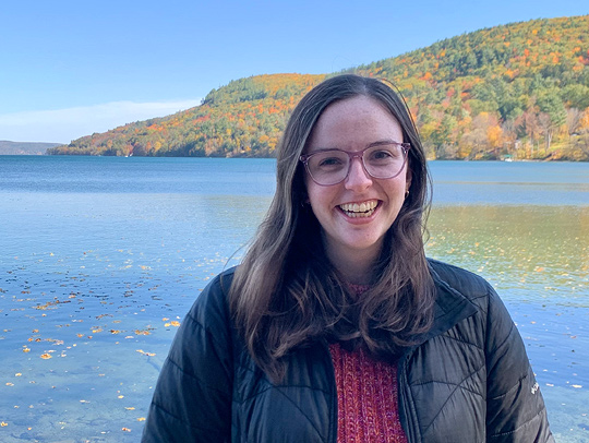 Victoria Whitley outside, in front of a river. Wooded hills with fall foliage is in the background.