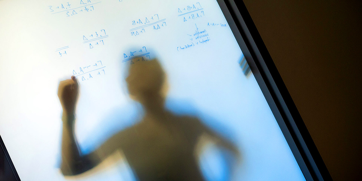 A person writing equations, seen in soft shadow from the opposite side of a frosted glassboard. Photo by John T. Consoli.
