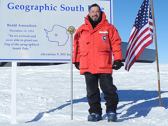 Gregory Sullivan at the monument marking the South Pole. He is wearing a red parka, black snowpants, and heavy boots.