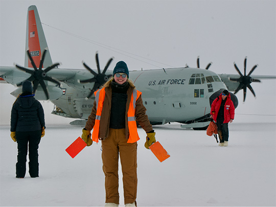 Rachel Procter Murphy at the South Pole wearing cold weather gear and a bright orange vest. A plane is on the ice behind her. Credit: E. Blaufuss.