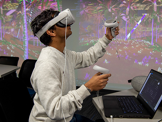 A student using a virtual reality headset and hand controllers, standing in front of a wall-sized screen showing the virtual environment. A laptop is on the dek in front of him. Credit: Lisa Helfert.