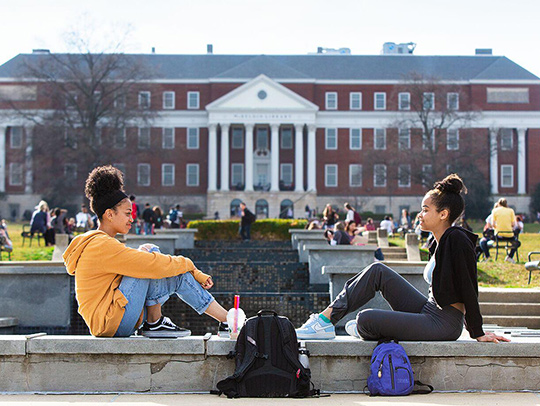 Two young women with backpacks sitting at the UMD mall fountain with the library in the background.