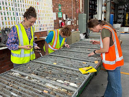The group visiting Mornington Core Storage, a library of drill core and rock samples. Three women in bright, reflective safety vests leaning over a large table covered in trays filled with rocks. A large chart is on the wall behind them. Image courtesy of Sarah Penniston-Dorland. The group visiting Mornington Core Storage, a library of drill core and rock samples. Three women in bright, reflective safety vests leaning over a large table covered in trays filled with rocks. A large chart is on the wall behind them. Image courtesy of Sarah Penniston-Dorland.
