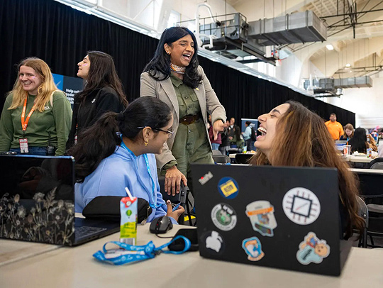 A group of women at Technica. Two are stitting at a table behind laptops, laughing with a mentor who is with them.