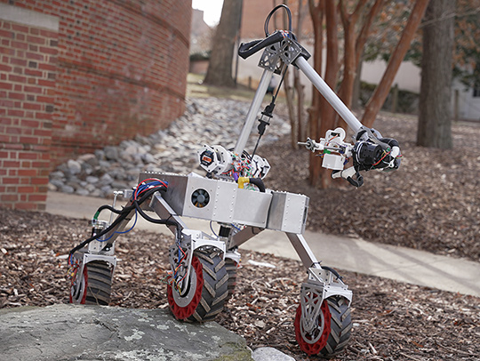 A small, 4-wheeled robot with a large arm mounted on its chassis traveling over rocks and mulch.
